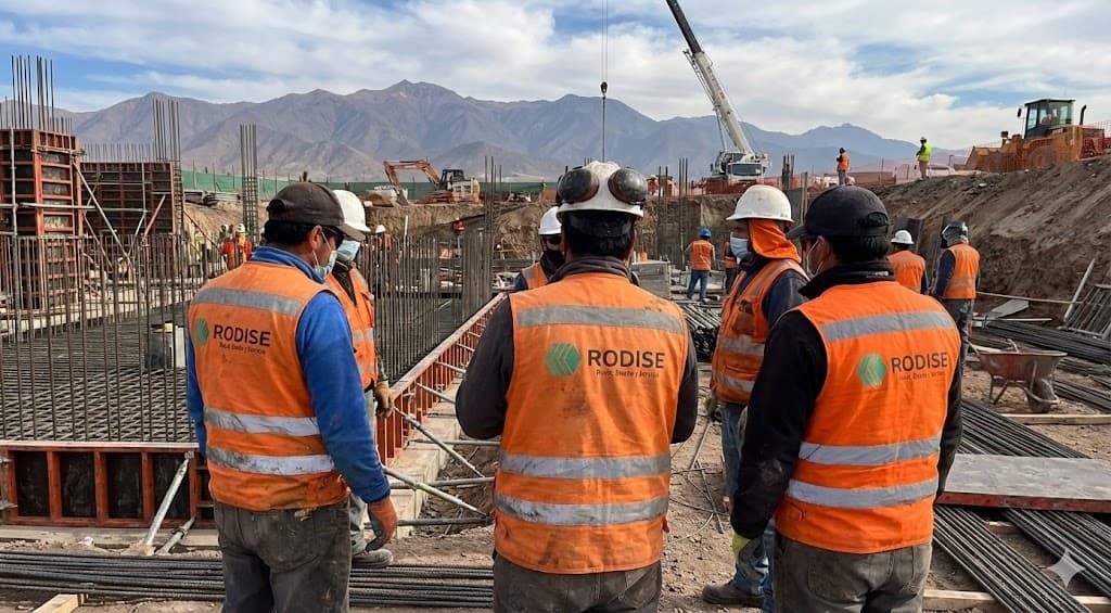 Construction workers in orange safety vests overlook a large foundation site with distant mountains.